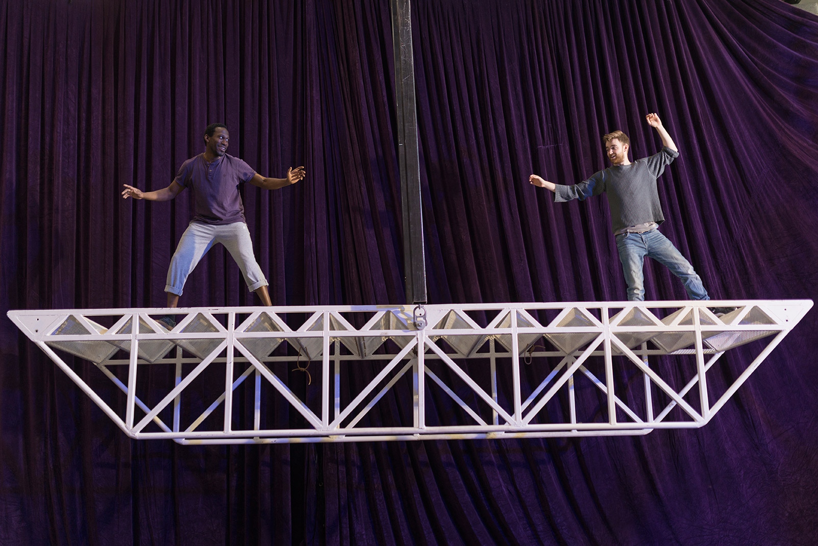 Two men stand on opposite ends of a 6 meters long bridge made of white metal. It is up in the air and acting as a balance, held in its centre by a large metal bar that allows it to balance to the left and to the right.
