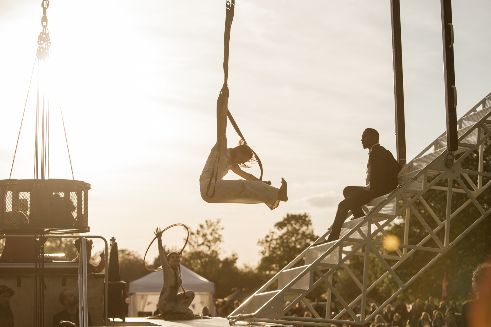 Aislinn swings on aerial straps above the Weighting show set. Behind her Nelly holds a hoop in the air and David sits on the staircase. They are all in silhouette as the sun lies low in the sky.