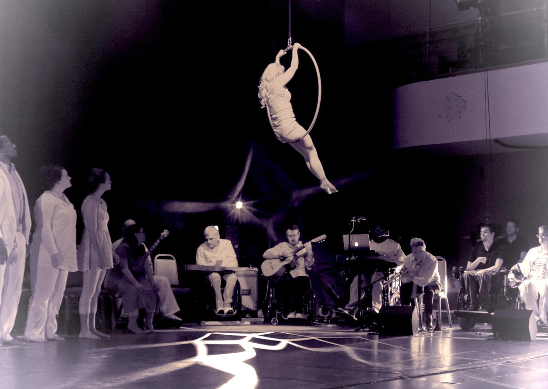 Karina sits on an aerial hoop as part of the Terry Riley 'In C' show. The rest of the cast and the musicians watch her from below.