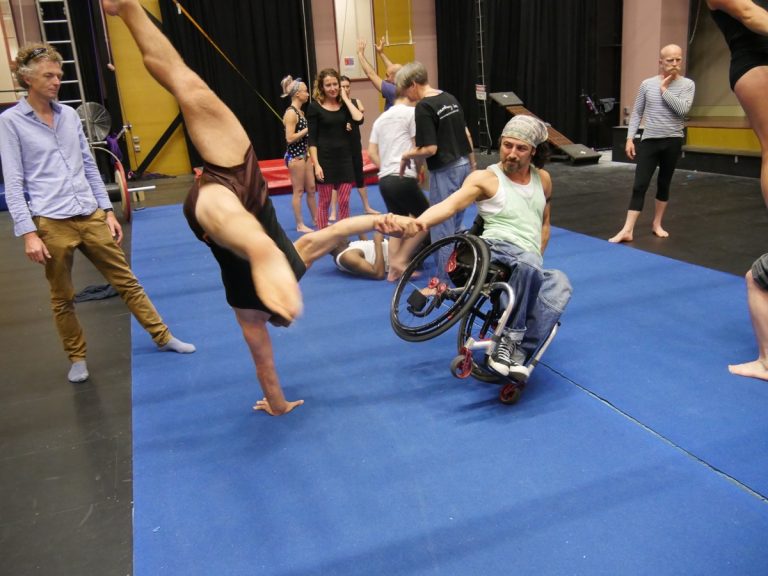 Dergin dances with his wheelchair, leaning over dynamically whilst holding the hand of an artist doing a handstand.