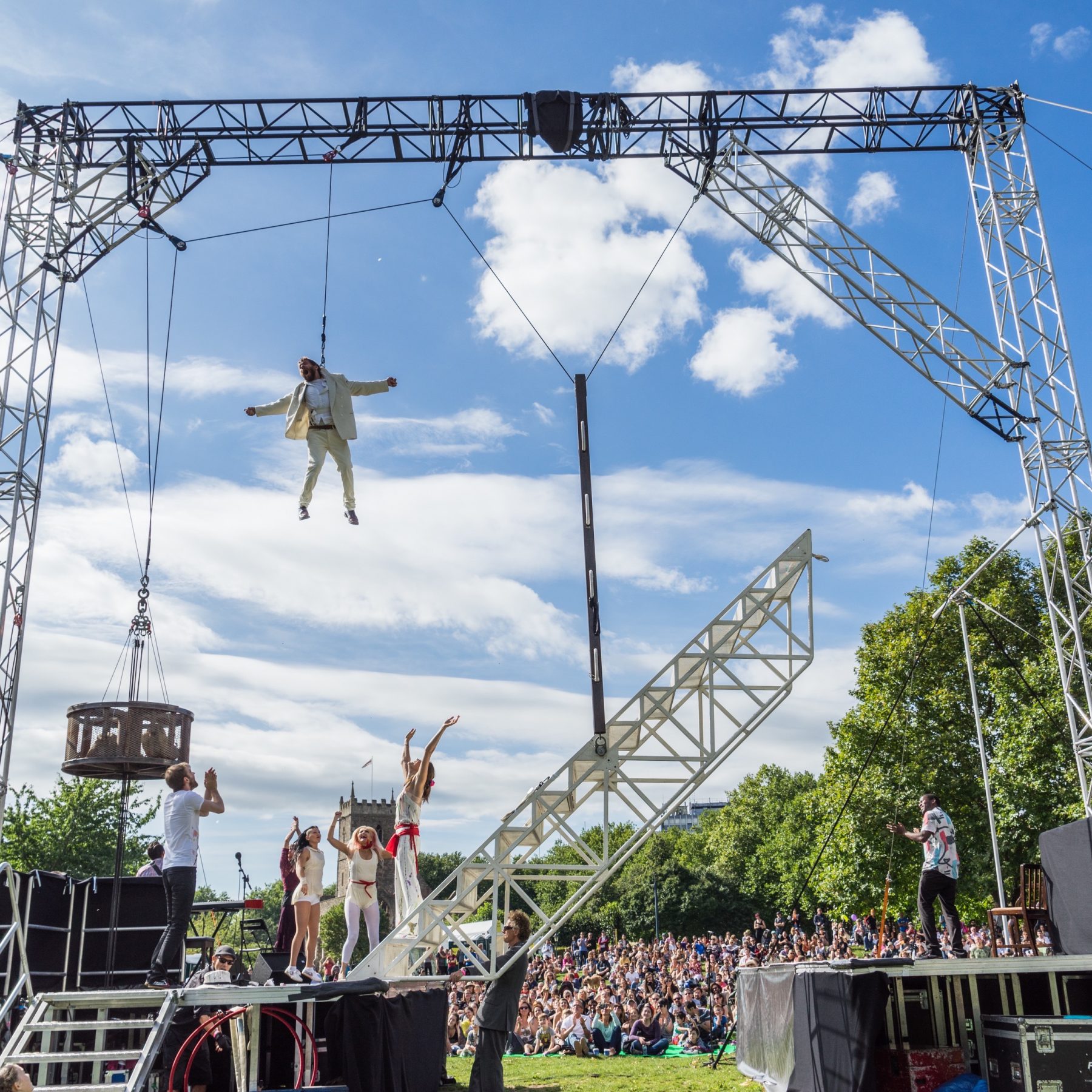 Weighting show in Castle Park. Jamie Beddard flying from large rig. Cast celebrate below, stretching arms up towards him. Large crowd watching show.