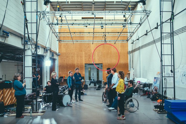 Extraordinary Bodies artists form 2 lines in the National Theatre Studios. A red aerial hoop hangs above their heads from circus rigging. Claire holds a microphone, Jonny is set up at the drums.