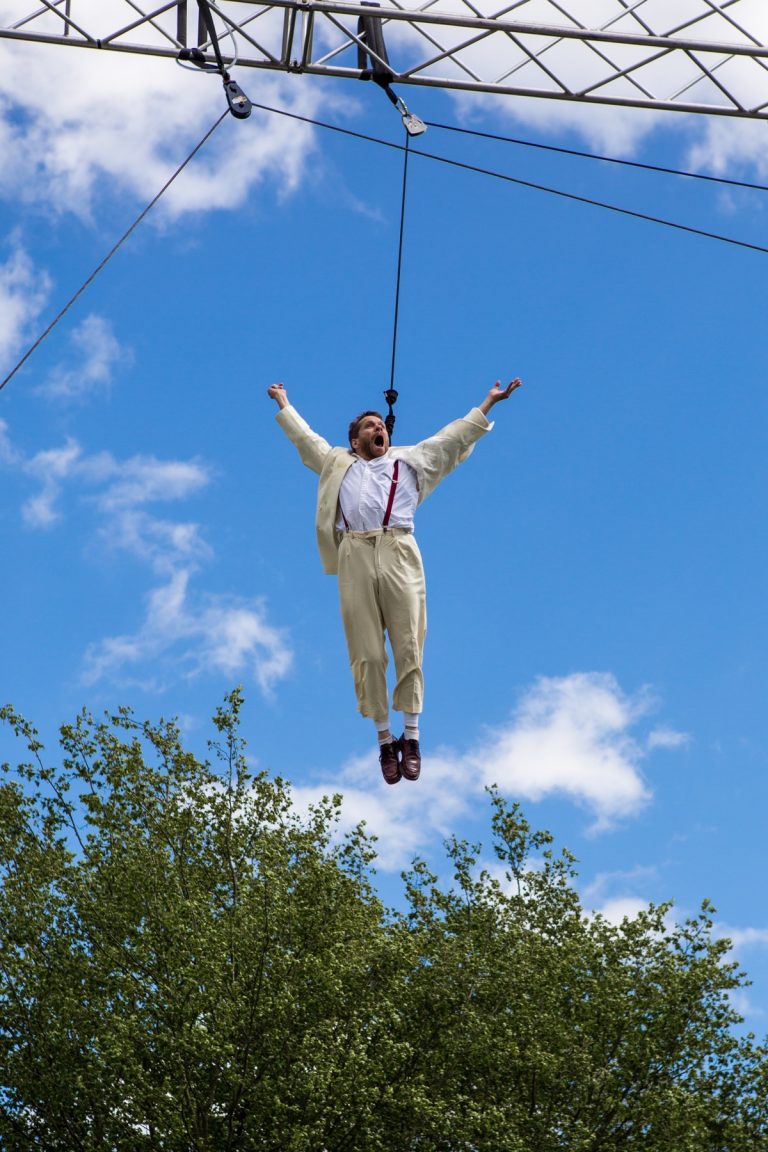 Jamie flys in the air on an aerial harness, his arms outstretched with an ecstatic expression on his face, a bright blue sky behind him.