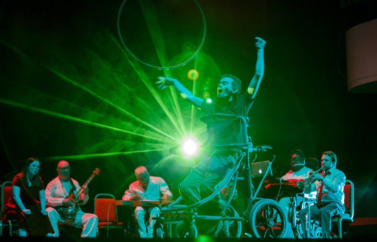 Dave Young sits in his wheelchair on stage, with green staging light bursting behind him. His arms are in the air and mouth wide open. The British Paraorchestra sit behind him playing instruments.