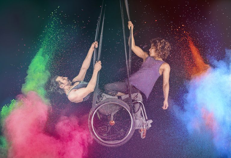 Suspended from the ceiling and surrounded by explosions of colourful chalk, two performers sit in a wheelchair, their muscly arms revealed