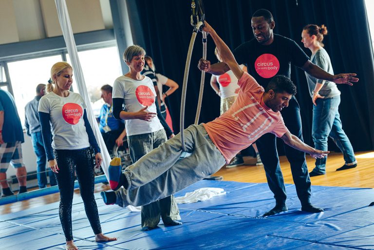 A man hangs off an aerial hoop in an Extraordinary Bodies workshop, three workshop leaders stand behind him wearing t-shirts saying 'Circus for Every Body' smiling and encouraging him