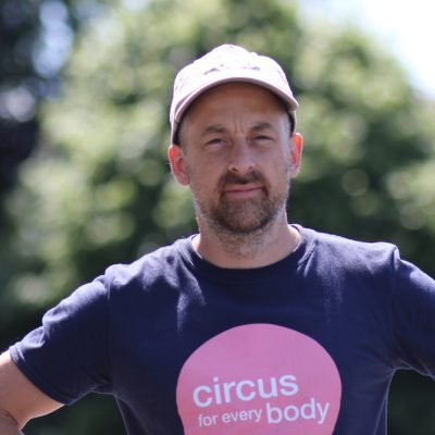 Headshot of Will Datson, a white man with dark stubble facial hair, wearing a 'circus for every body' tshirt and a cap.