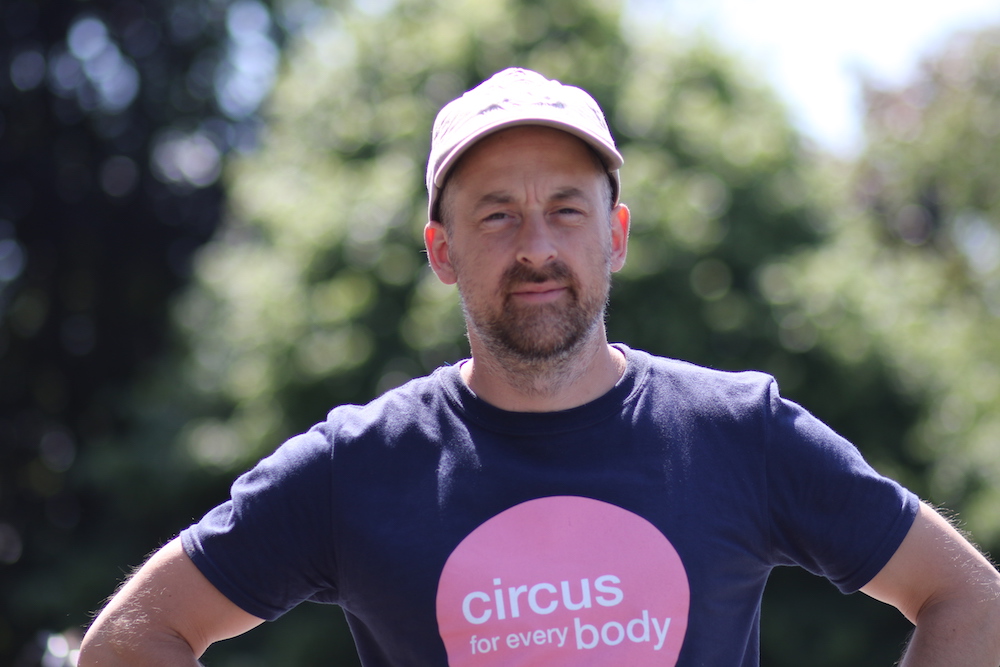 Headshot of Will Datson, a white man with dark stubble facial hair, wearing a 'circus for every body' tshirt and a cap.