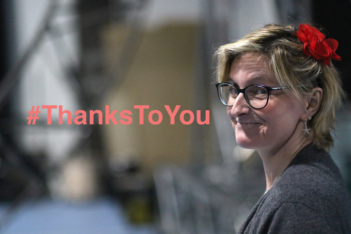 Co Artistic Director Claire Hodgson turns to the camera smiling, a red flower in her blonde hair. Text reads #ThanksToYou