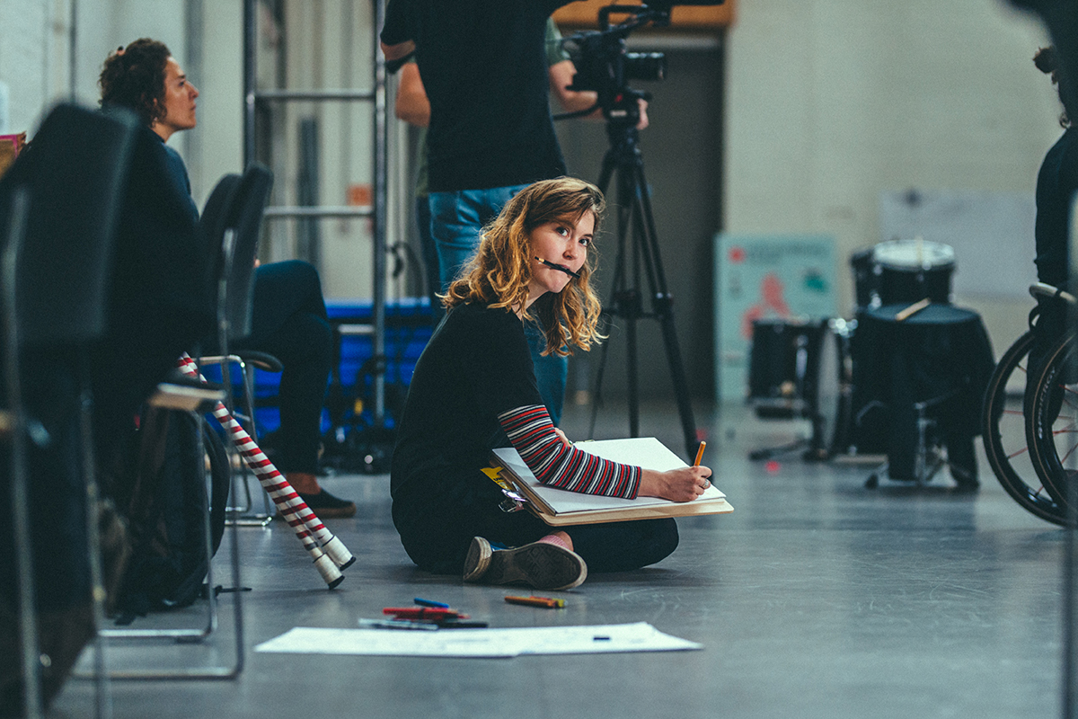 A female illustrator sits on the floor of the rehearsal studio, one pen in her hand and the other in her mouth, sketching