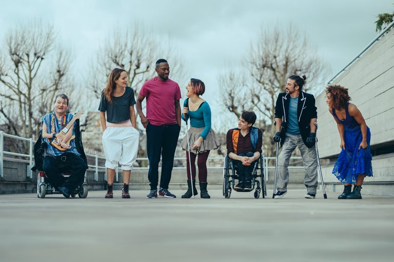 Seven artists line up outside standing on grey concrete. They wear colourful clothes and look towards each other laughing. Two of the cast are in wheelchairs, one holds a stick and one holds crutches. They are a mix of male female, black, white and mixed race, disabled non-disabled.