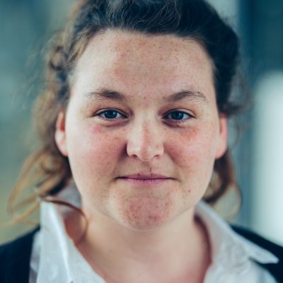 Eilis Davis, smiling to camera - Eilis has long, wavy, dark brown hair tied up. A gentle smile, lots of freckles and deep blue eyes.
