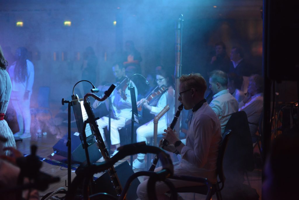 The British Paraorchestra musicians, playing at Birmingham Town Hall. The lighting is dark and moody. A soft spotlight illuminates musicians playing violins and a clarinet.