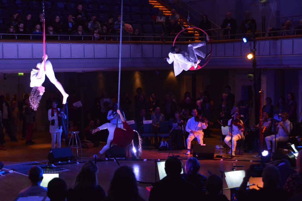 Wide shot of Birmingham Town Hall, the musicians seated in a large circle around the room, creating a centre stage for the aerial artists. One girl suspended upside down in a red hoop, another man perches with his legs high, sat in another red hoop. A second girl is flowing down a rope, also suspended from the ceiling