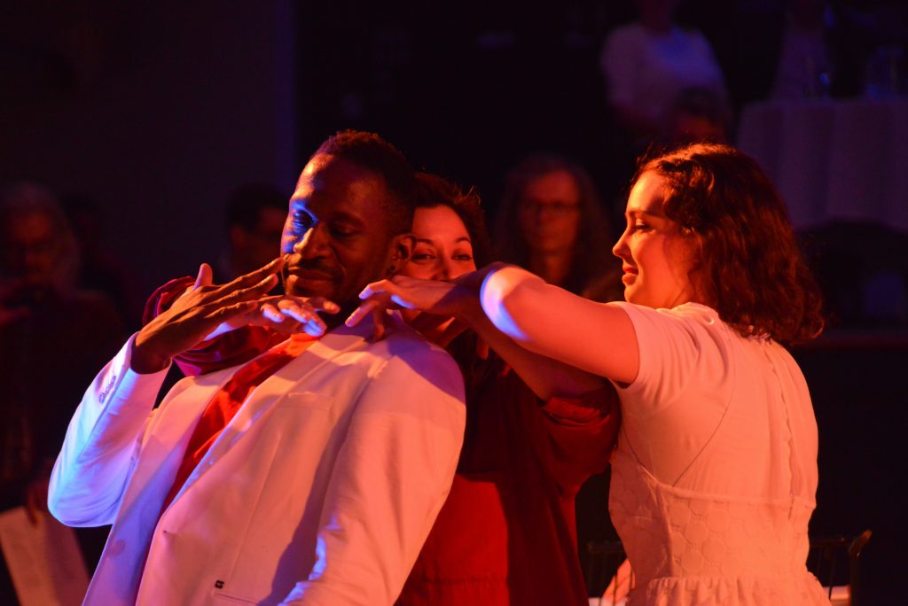 Three artists are intertwined with arms and hands, illuminated by a soft red light. They appear to be bouncing or moving gently to a pulse or beat of the music.
