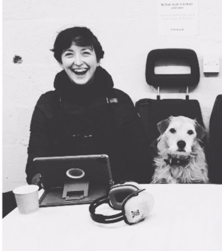 Photograph of Georgina, laughing to camera. She's a white woman, with short dark hair. She's sat next to a small scruffy light haired dog.