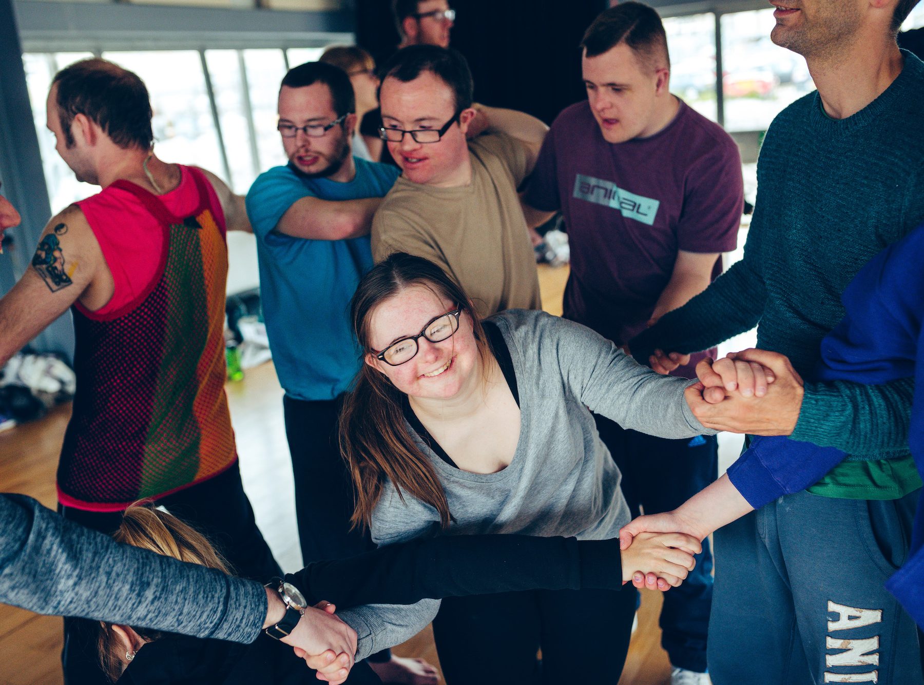 A group of young people are tangled in a group, holding hands, with arms intertwined. A young girl with Jess, a young girl with Downs Syndrome is at the front of the group, beaming with a big smile.