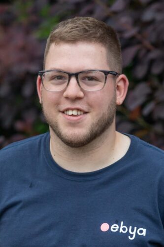 Headshot of Harry. He is a white man in his twenties with short brown hair, black glasses and stubble beard. He is smiling brightly at the camera.
