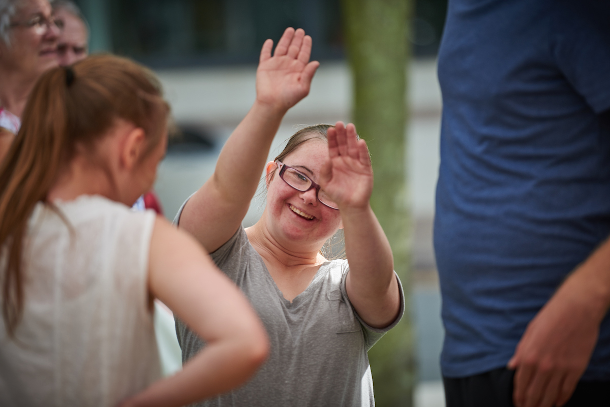 Jess, a young girl with Downs Syndrome, waving to camera with both arms in the air, smiling.