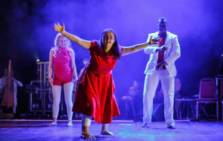 Extraordinary Bodies performing on stage with British Paraorchestera; a learning disabled woman stands centre stage, lunging forward with her arms outstretched. She wears a long, bright red dress. The stage lighting is a moody purple. Two more artists move in the background.