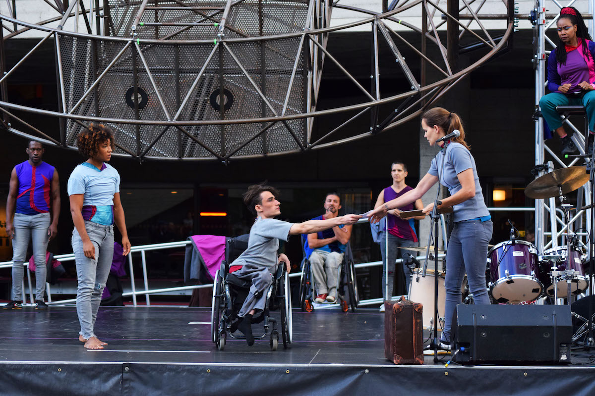 Extraordinary Bodies perform What Am I Worth? at London's National Theatre Riverstage Festival. Jonny is centre stage in his wheelchair; reaching out to Aislinn's hand to take a piece of paper. The rest of the artists look on with concerned expressions and frowns.