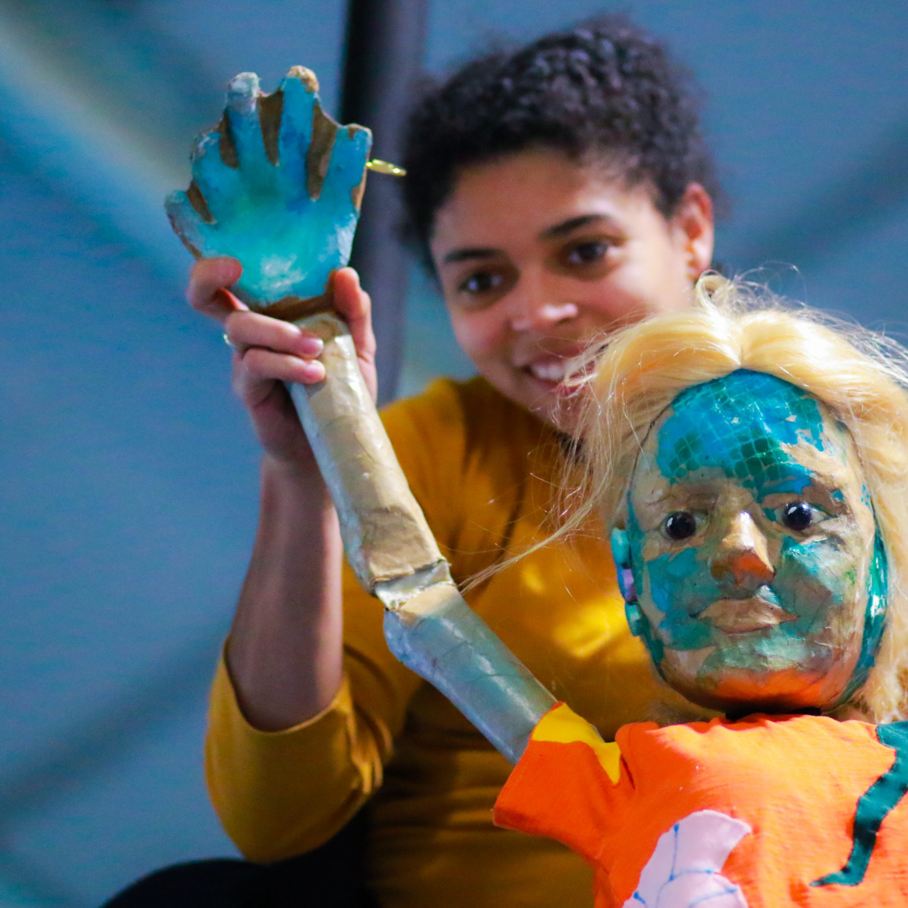 A young mixed-race woman is smiling and holding up a puppet of a girl with blond hair, an orange top and fish-like turquoise scales of her face.