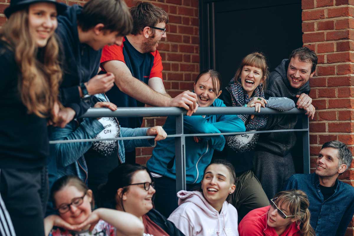 A group of disabled and non-disabled young people and adults laughing together, outside a red brick building, interacting with one another.