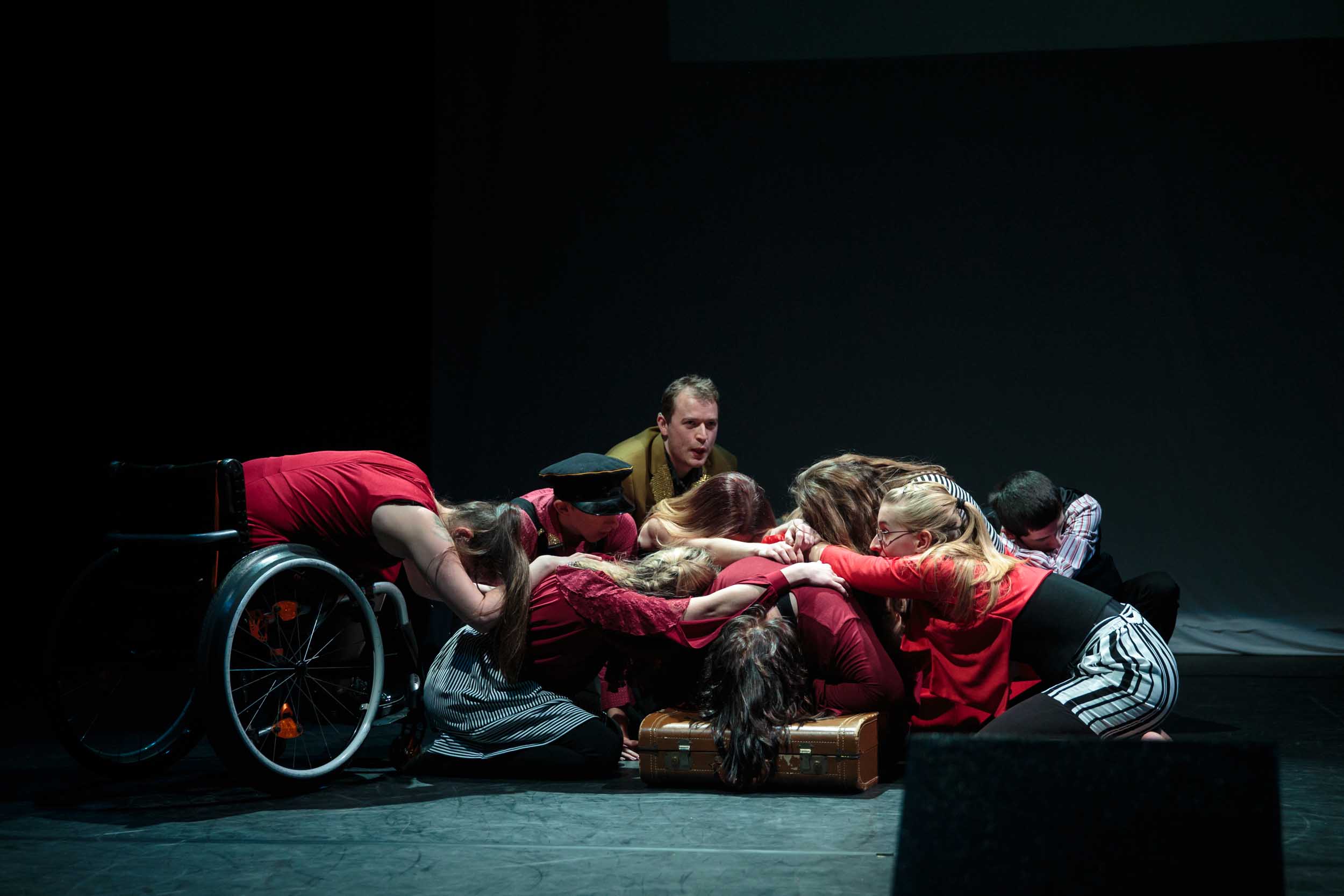 A group of young disabled and non-disabled people grouped together in the centre of a dimly lit stage. They are all sitting or bending their bodies towards the ground, holding onto each other.