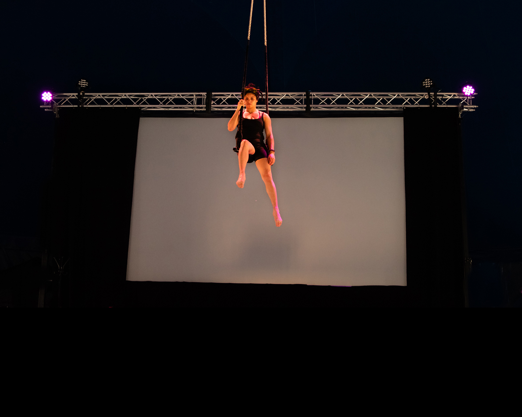 A woman in her twenties with light brown skin and afro hair twisted into a half-up half-down style sits on a trapeze, flying in the air. One of her legs is bent and pointing towards us. Behind her, steal rigging and purple lights and a white screen.