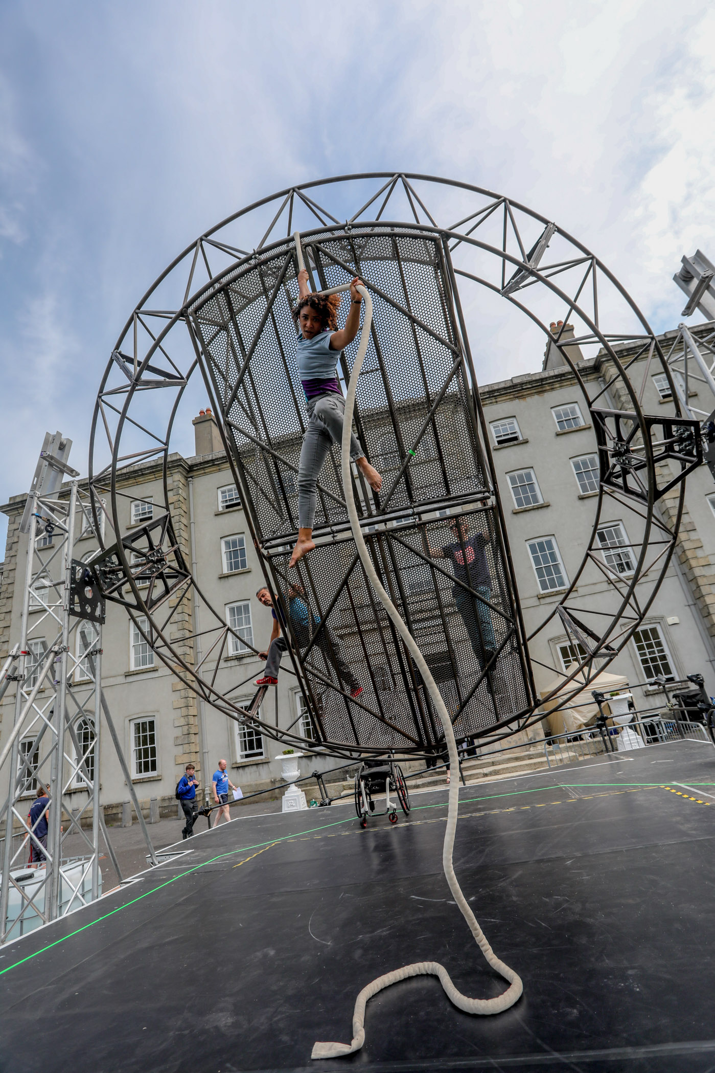 A giant steel wheel on an outdoors circus stage. The wheel is held to the side by two axis that allow it to tilt forward and back. A woman, short and slim with black afro hair, holds herself up in the air with a circus rope that is attached to the part of the wheel that points towards the sky at the moment when the photo is taken.