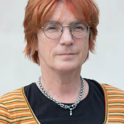 Headshot of Abi, a white woman with short hair dyed orange, round and thin-rimmed glasses. She has a nose ring and wears a thick chain around her neck.