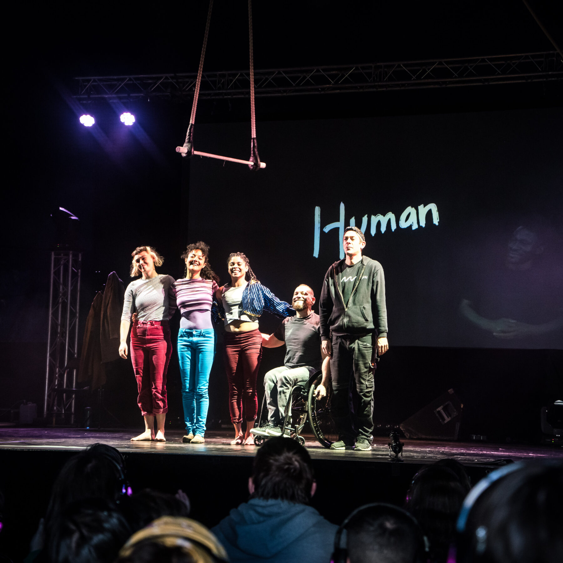 An indoors stage. The space is dark apart from two purple spotlights at the back. Performers stand in line and prepare to bow to the audience, which can be seen in the foreground. Behind them, a screen shows a title in white handwriting: "Human" and a man who BSL interprets. Above, a trapeze can be seen hanging above the performs.