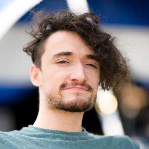 Jonny: a white man in his twenties with thick brown hair flicked to the side and a stubble beard. He smiles brightly at the camera.
