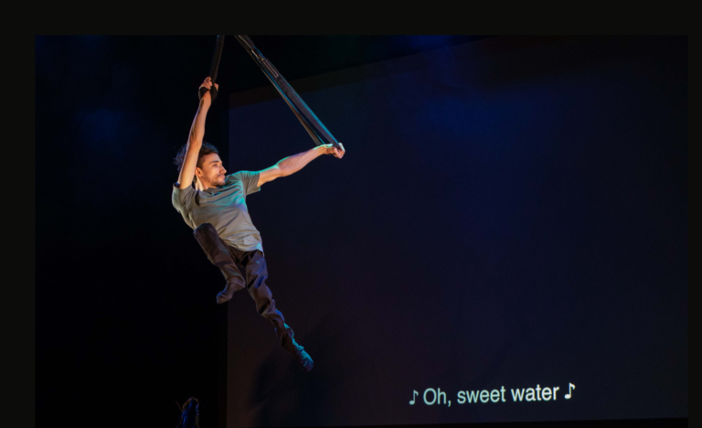 A white man in his twenties in the air above the the stage by holding onto black straps. He wears a grey T-shirt and black trousers. His thin legs dangle below him as he flies, looking strong. In the background, white text on a black screen tells us that a song is playing in this moment, with the following lyrics: "Oh, sweet water".