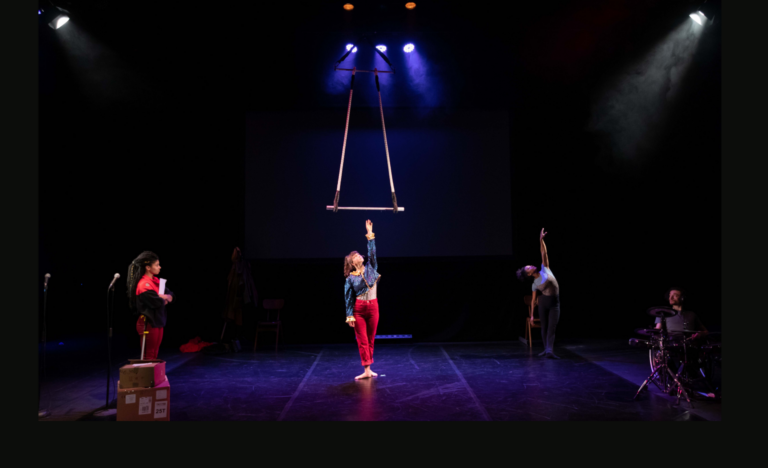 Three performers on a dimly lit bare stage, purple spotlights above them. In the centre, a white woman in her twenties stands with one arm raised up to a trapeze that hangs from above. She wears a blue and gold sparkly top and red trousers. On the left, a mixed-race woman in her twenties wears a DPD delivery outfit - black and red fleece top and red trousers. She looks at the performer in the centre. On the right, a mixed-race woman stands with one arm raised up, bending backwards gracefully. She wears a leotard and grey leggings.
