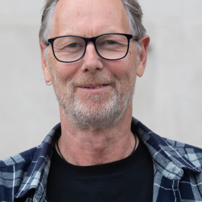 Headshot of James: A middle-aged man with grey hair, a white stubble beard, black-framed glasses. He is wearing a blue and white checked shirt and is smiling softly at the camera.