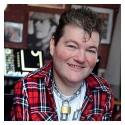 John: a white, Irish male. He has dark hair with a Kiss curl on the forehead. He always wears short sleeved t-shirts, and is an electric wheelchair user with studded seatbelt. He wears a padlock on a chain as a necklace. He is smiling brightly at the camera.