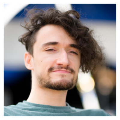 Jonny: a white male in his late twenties with a stubbly beard and a large floof of dark hair on top. He uses a manual wheelchair with a stout body and long arms. He smiles brightly at the camera.