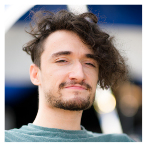 Jonny: a white male in his late twenties with a stubbly beard and a large floof of dark hair on top. He uses a manual wheelchair with a stout body and long arms. He smiles brightly at the camera.