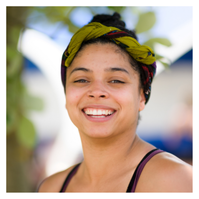 Rebecca: a woman in her twenties with light brown skin and beautifully untameable afro hair. She wears a green and red headwrap twisted around her head. She is smiling brightly at the camera.