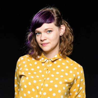 Headshot of Monika, a white woman in her twenties with shoulder-length wavy brown hair, blue eyes and a yellow shirt with white polka dots.