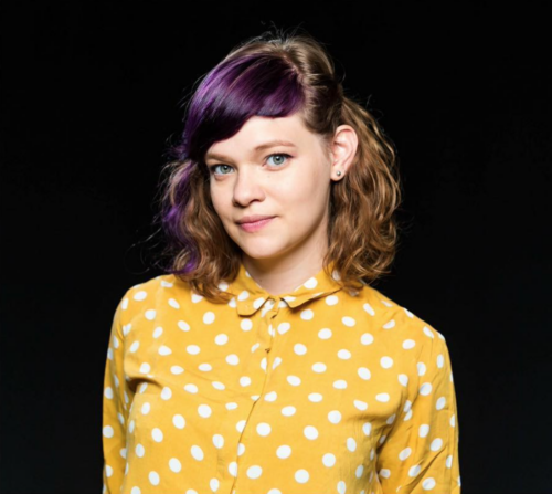 Headshot of Monika, a white woman in her twenties with shoulder-length wavy brown hair, blue eyes and a yellow shirt with white polka dots.