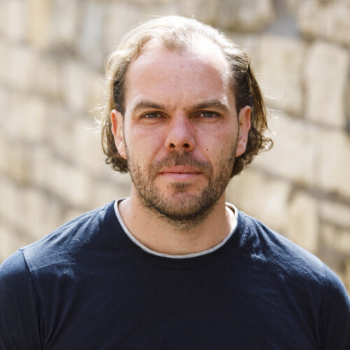 Steven: a white man with ear-length, wavy, brown hair and a brown stubble beard, wearing a dark blue t-shirt. He is standing outside, smiling softly at the camera, the sun touching the top of his head.