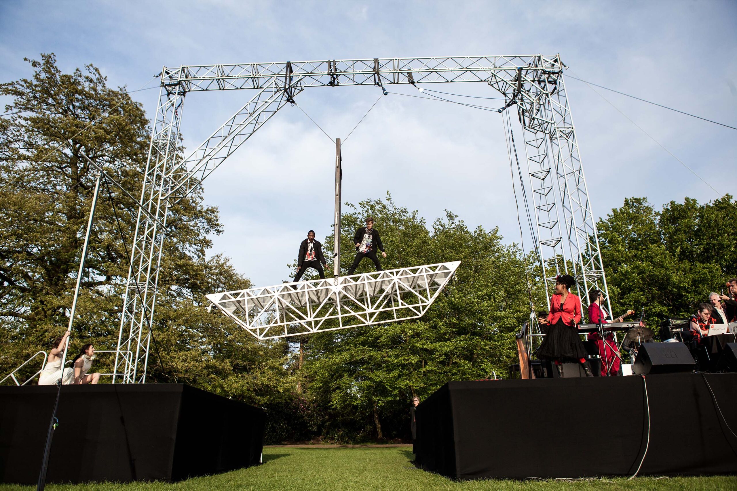 An outdoors circus stage with tall steel rigging. Two men stand on opposite ends of a 6 meters long bridge made of white metal. It is up in the air and acting as a balance, held in its centre by a large metal bar that allows it to balance to the left and to the right.