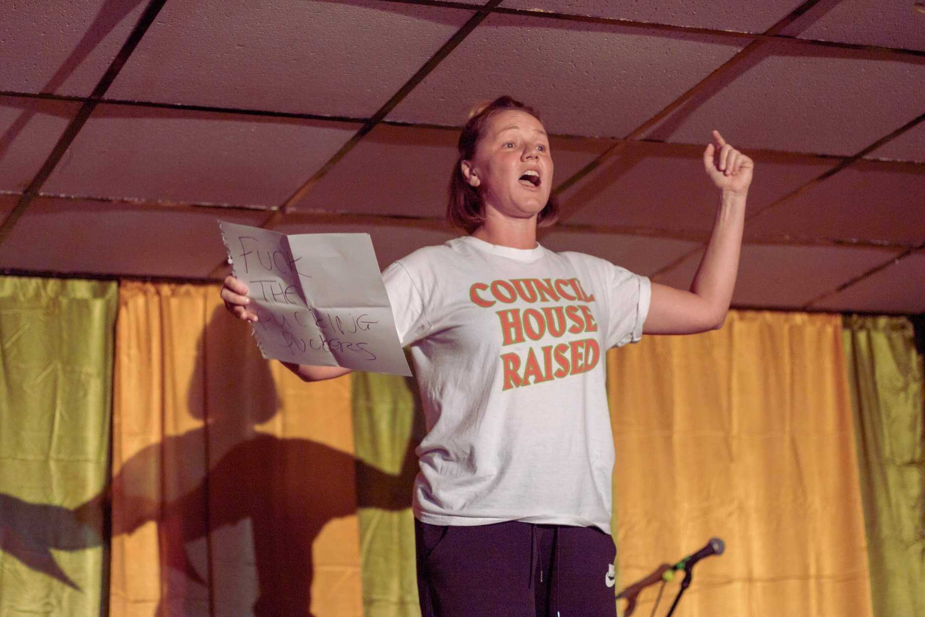 Jodie rehearsing. She is standing in a low ceiling room, yellow and pistachio green curtains behind her. She wears a white t-shirt with all caps orange letters on it that read: "Council house raised". She holds a white sheet of paper with one hand, on which can be seen the words: "FUCK THE FUCKING FUCKERS". Her other hand is raised up in the air in protest and her faces suggests shouting.