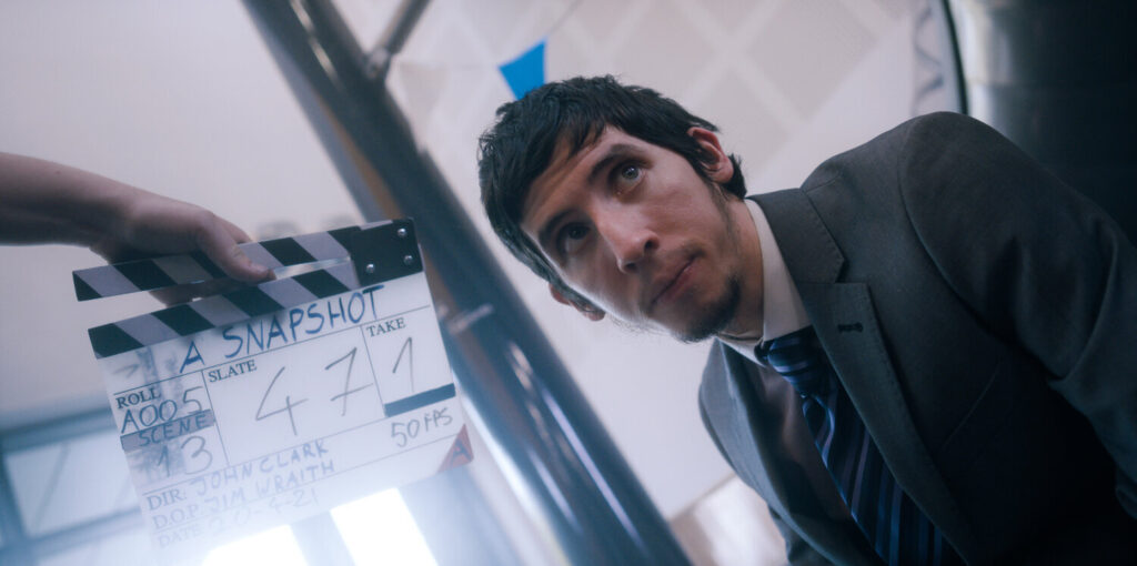 A young white man in his twenties with short black hair and a soft beard. He is wearing a grey suit with a black and blue striped tie. He is looking away from the camera. To his left, a hand holds a film clapboard. Text on it reads: "A SNAPSHOT, Slate 47, Take 1. Director John Clark".