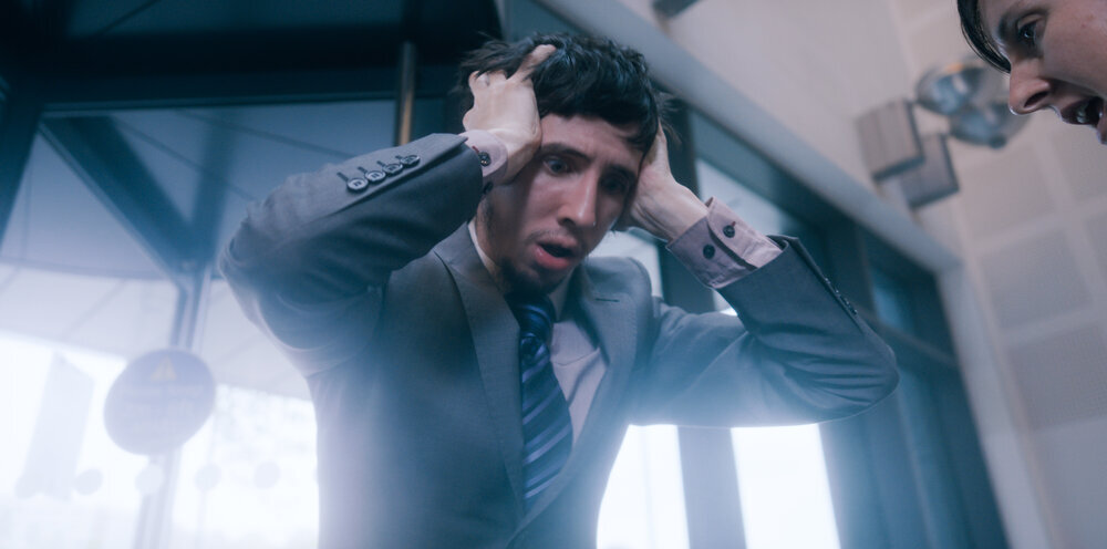 Josh, a white man in his twenties with short black hair and a soft beard, holds his head in his hands. He looks deeply distressed and his mouth is open. He looks like he is in a building's lobby. In the top right corner, a small part of a person's face can be seen. They look angry.