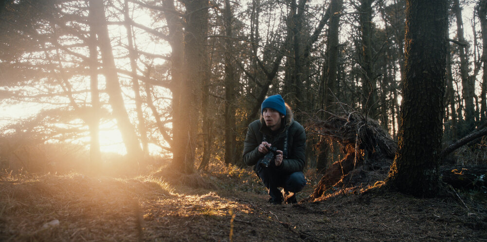 Josh, a white man in his twenties, crouches in a forest. He is wearing a blue beanie hat and a warm jacket. He is holding a camera and looking at the camera the captures his image. Warm sunset light comes through the trees and illuminate the ground softly.