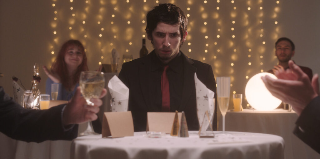 Josh, a white man in his twenties with short black hair and a soft beard, is sitting at a table. The environment looks like a party one - perhaps a wedding. There are fairy lights hanging from the wall in the back. There is a glass of sparkling, name cards and glasses with start-patterned napkins on his table. In the foreground, a hand holding a glass of fizzy alcohol and another pair of hands clapping. In the background, a female-presenting person and a male-presenting person clap.