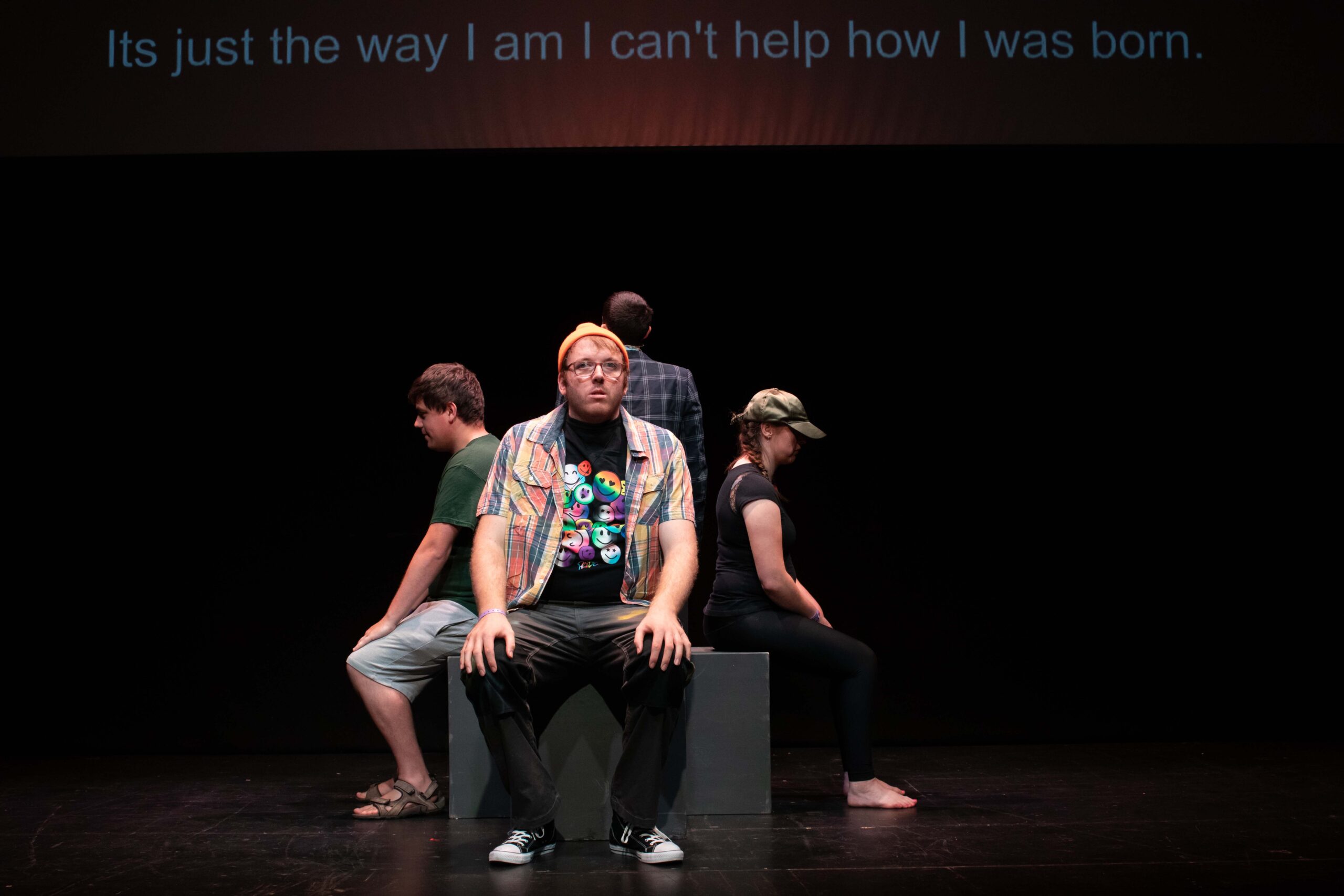 A group of young people sitting on a black block on a dimly lit stage. Each one of them sits on a different side of the block, back to back, looking pensive. Behind them, captions on a screen read: "It's just the way I am I I can't help how I was born."